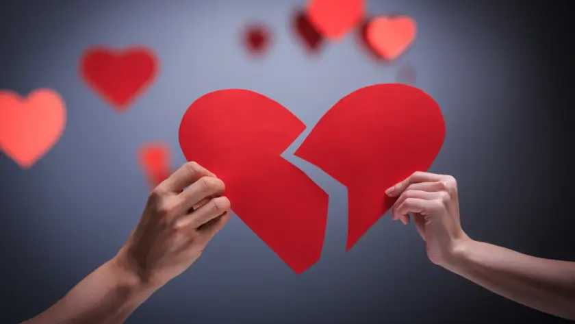 Two hands holding halves of broken red heart with blurred hearts in background