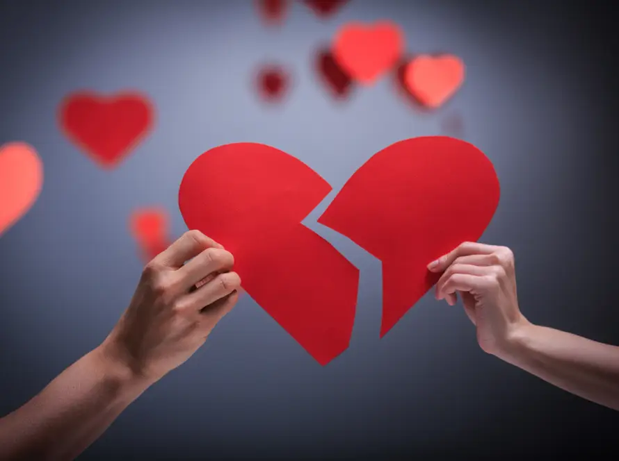 Two hands holding halves of broken red heart with blurred hearts in background